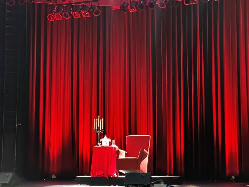 Bühne mit rotem Vorhang, davor ein roter Sessel und ein Tisch mit roter Decke und Kronleuchter.
